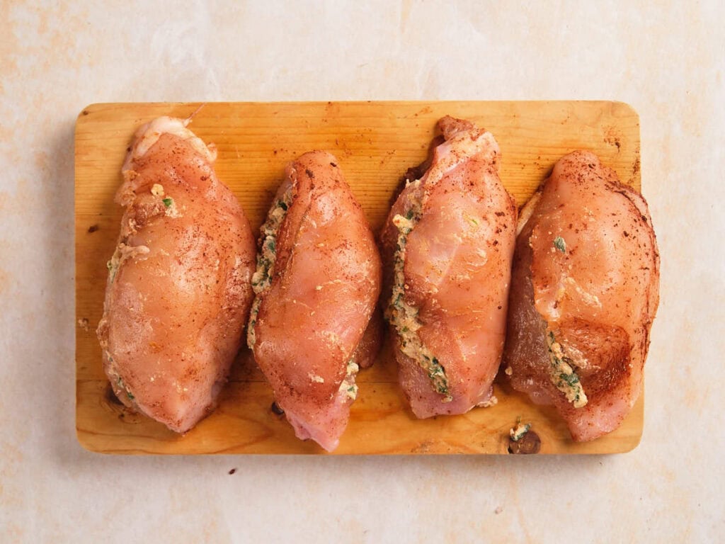 Four raw, seasoned chicken breasts stuffed with a cheese and herb mixture are arranged in a row on a wooden cutting board.