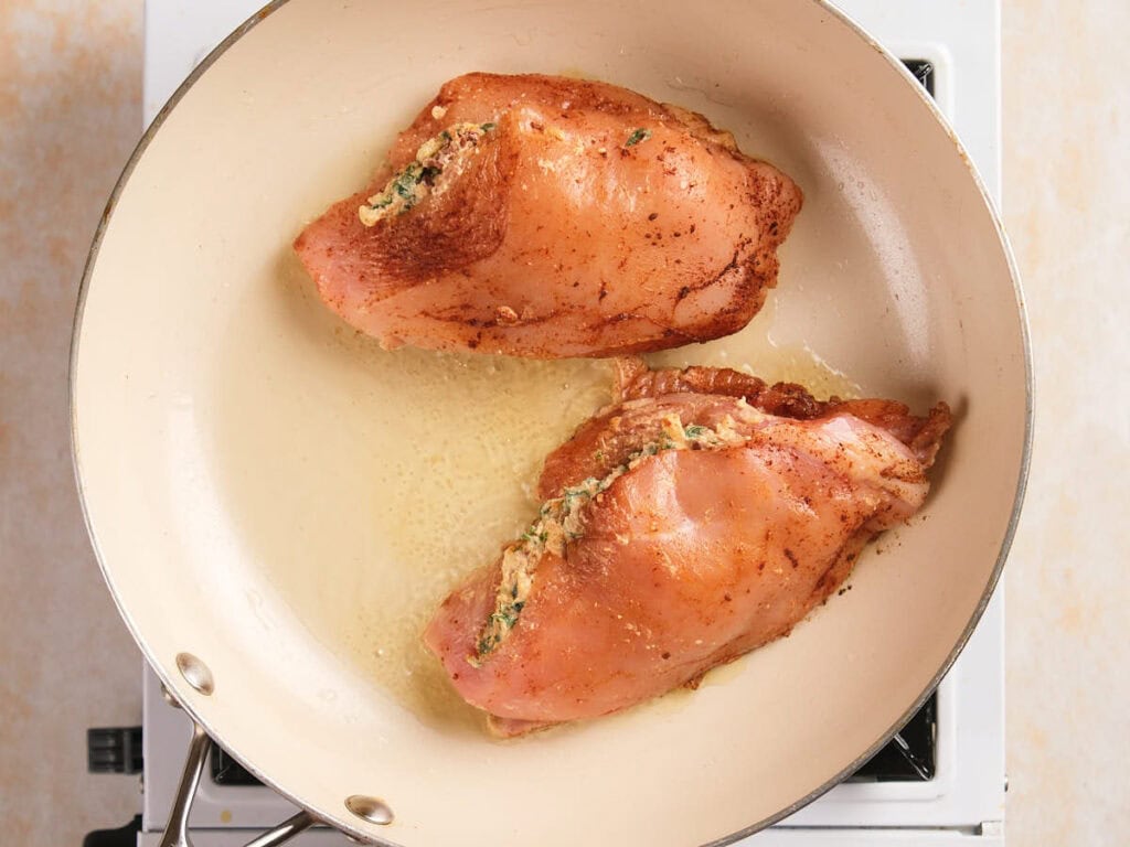 Two stuffed chicken breasts are being cooked in a light-colored frying pan on a stovetop.