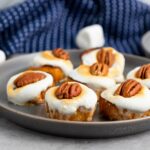 Six toasted marshmallow-topped mini desserts, each garnished with a pecan, are arranged on a gray plate with a blue cloth in the background.