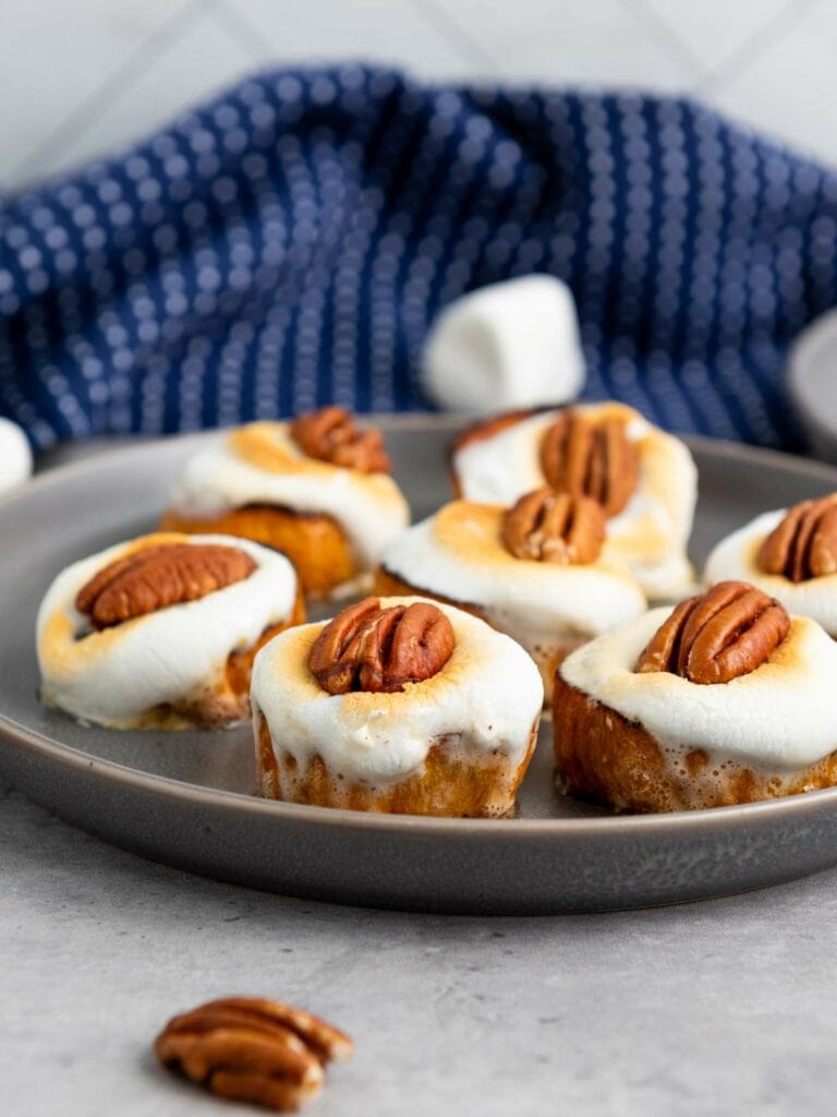 Six toasted marshmallow-topped mini desserts, each garnished with a pecan, are arranged on a gray plate with a blue cloth in the background.