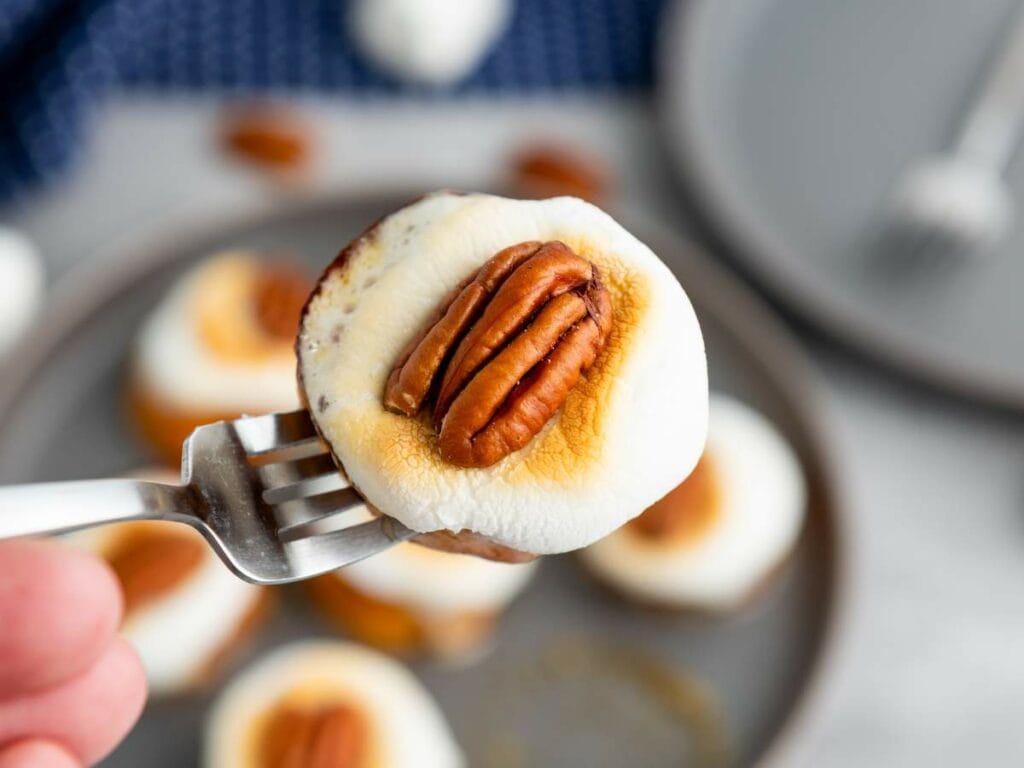 A fork holds a slice of sweet potato topped with a toasted marshmallow and a whole pecan, with more on a plate in the background.