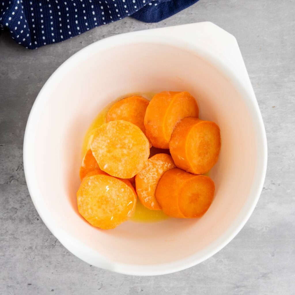Sliced sweet potatoes in a white mixing bowl with melted butter, on a gray countertop next to a blue cloth.