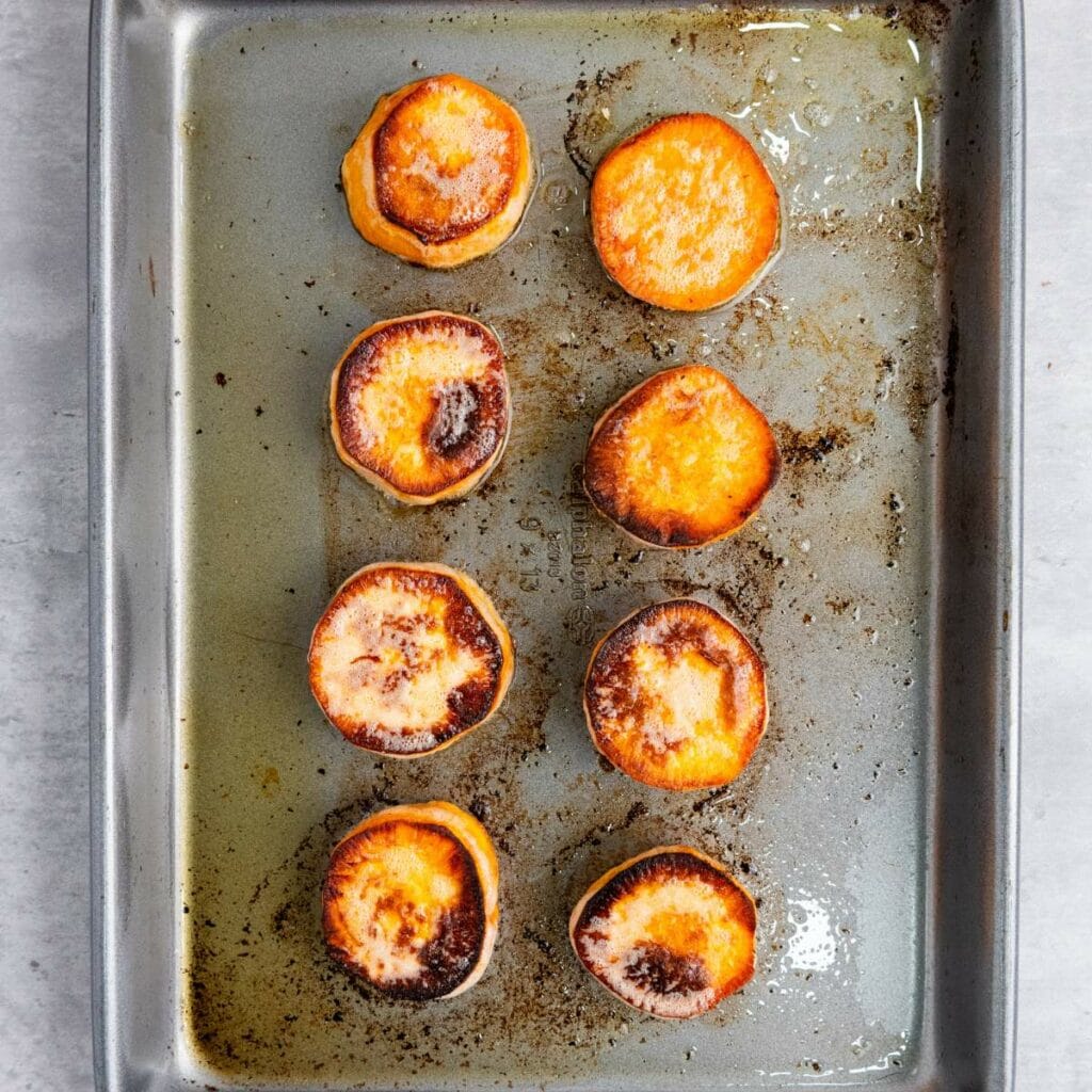 Eight roasted sweet potato slices on a metal baking sheet, some with browned edges and slight charring.