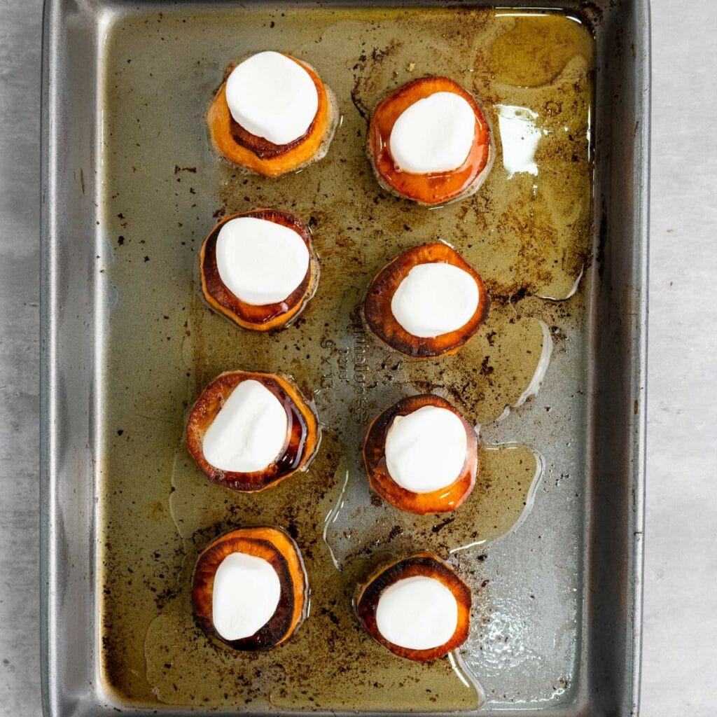 Eight baked sweet potato rounds topped with marshmallows are arranged on a greased metal baking sheet.