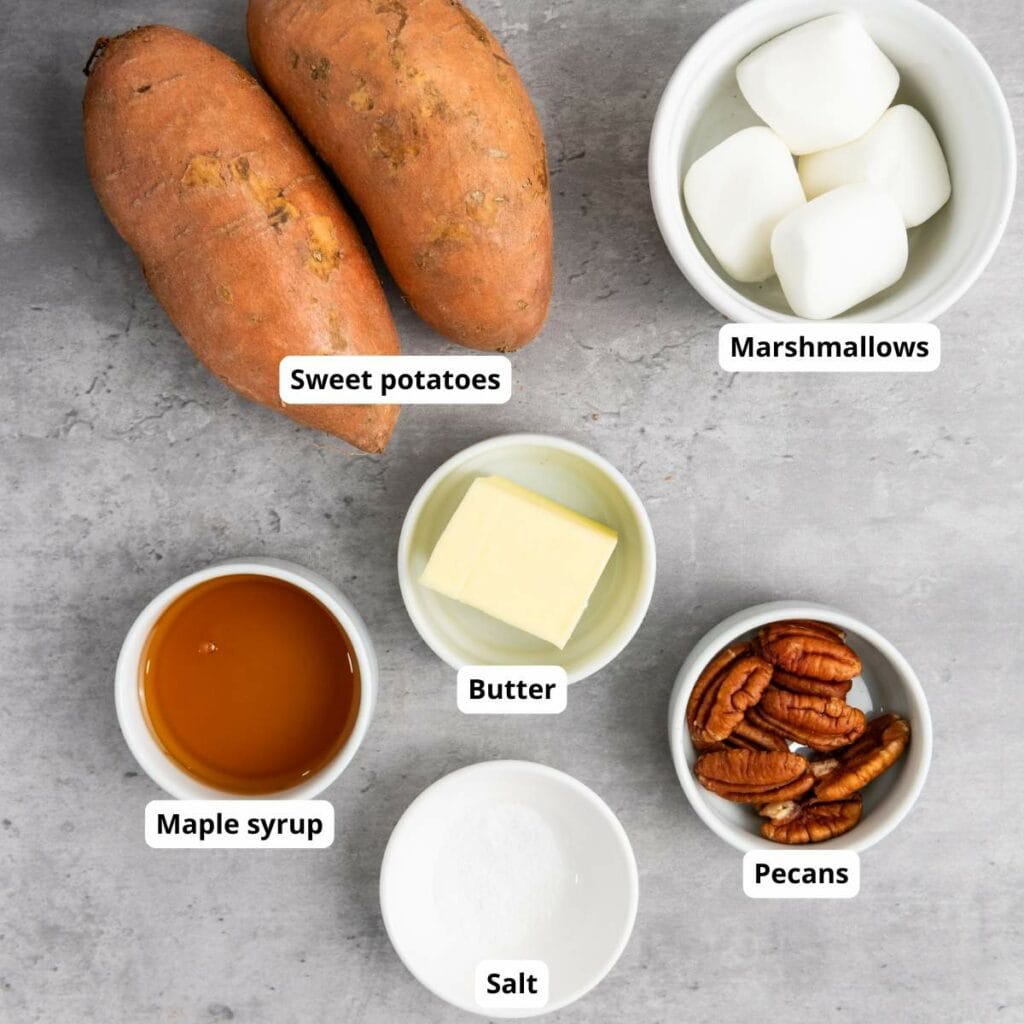 Labeled ingredients on a gray surface: sweet potatoes, marshmallows, butter, maple syrup, salt, and pecans in separate bowls.