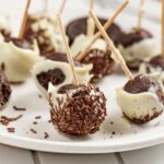 Chocolate cake pops coated in white chocolate and chocolate sprinkles, each with a stick, arranged on a white plate.