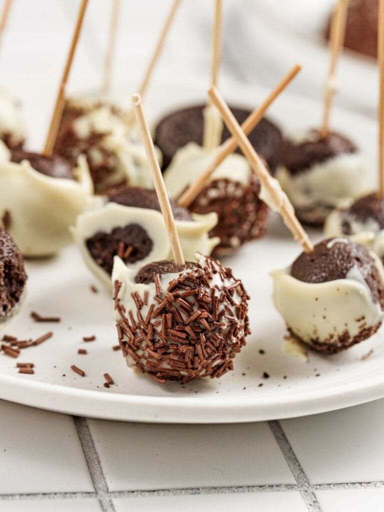 Chocolate cake pops coated in white chocolate and chocolate sprinkles, each with a stick, arranged on a white plate.