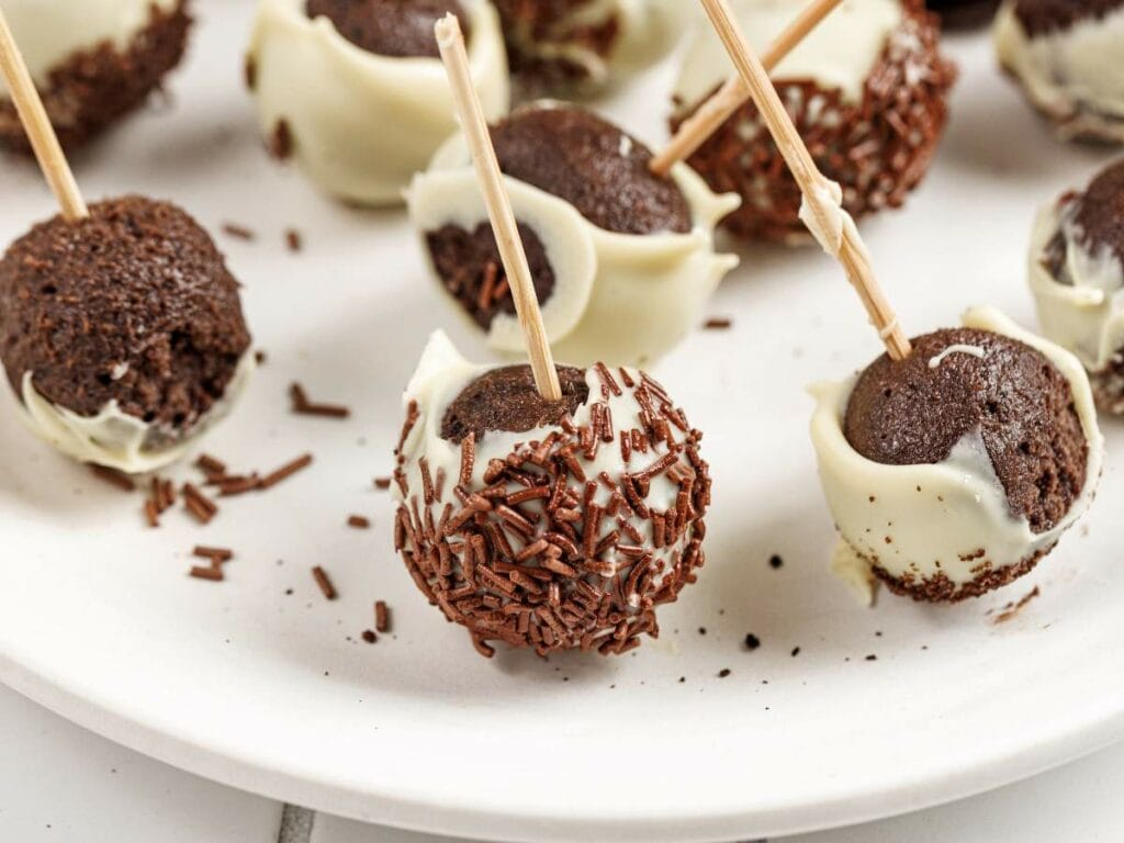Chocolate cake pops with white chocolate coating and chocolate sprinkles, each on a stick, arranged on a white plate.