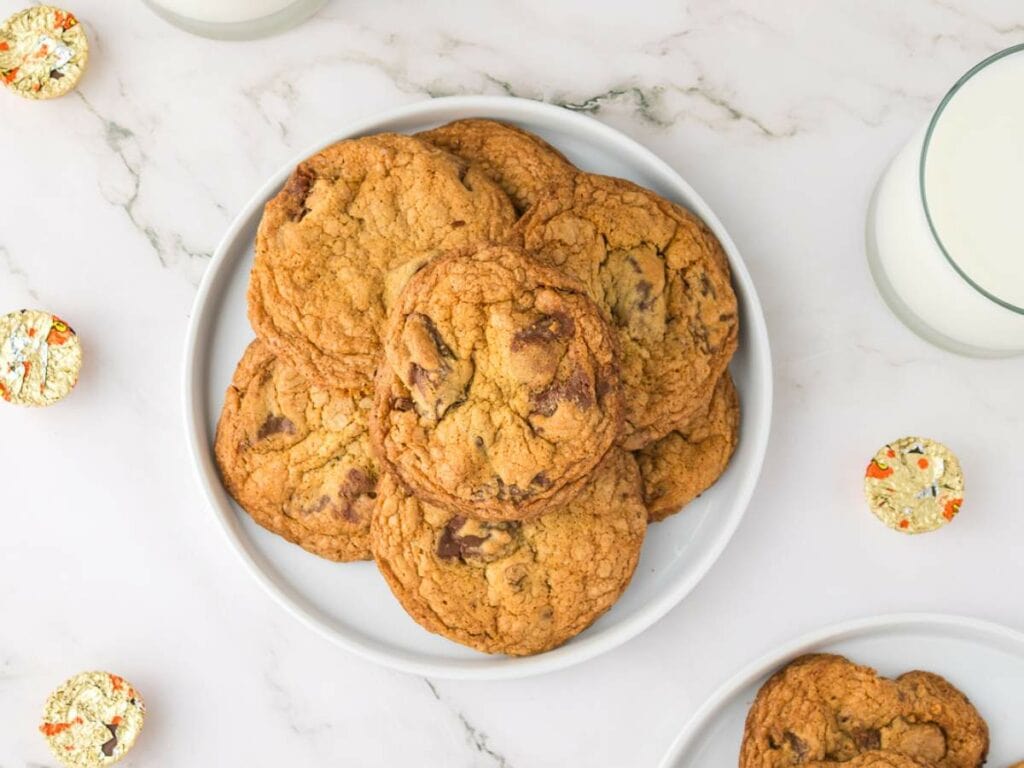 A white plate with peanut butter cup cookies sits on a marble surface, surrounded by glasses of milk and wrapped candy pieces.