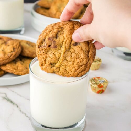 A hand is dipping a peanut butter cup cookie into a glass of milk. Plates of cookies and glasses of milk are in the background on a white surface.