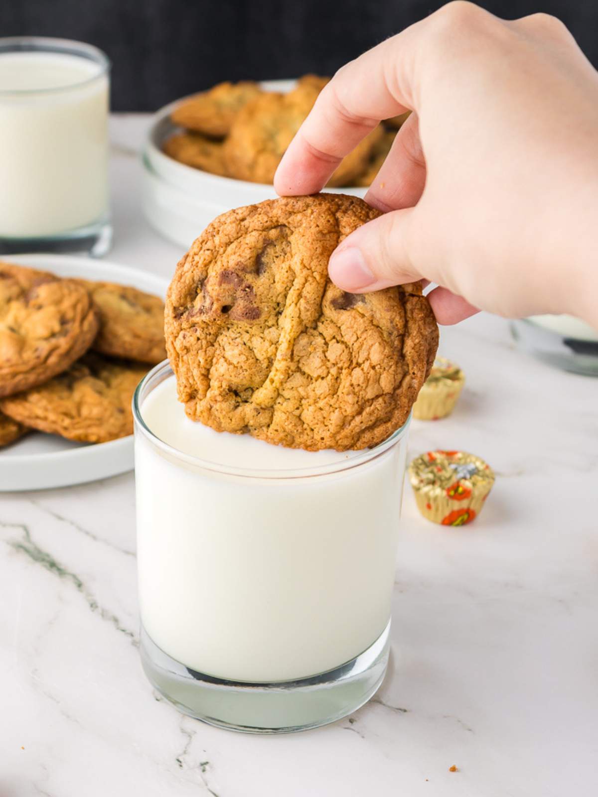 A hand is dipping a peanut butter cup cookie into a glass of milk. Plates of cookies and glasses of milk are in the background on a white surface.