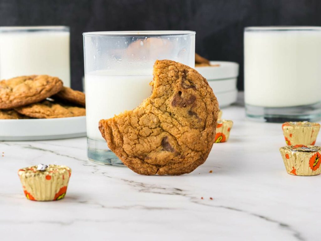 A peanut butter cup cookies with a bite taken out rests against a glass of milk, with peanut butter cups and more cookies on plates in the background.