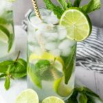 A glass of iced mojito garnished with lime slices and fresh mint, with a striped straw, sits on a white surface next to extra lime wedges and mint sprigs.