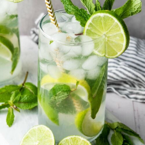 A glass of iced mojito garnished with lime slices and fresh mint, with a striped straw, sits on a white surface next to extra lime wedges and mint sprigs.