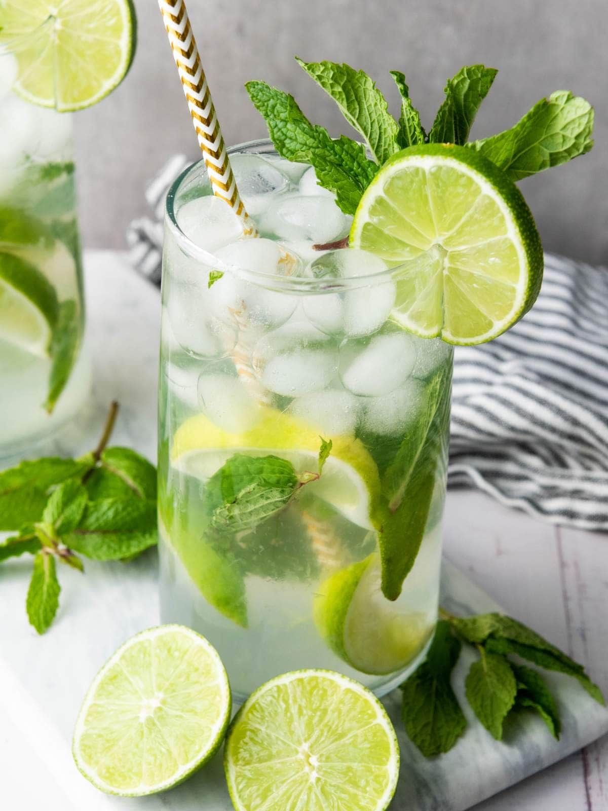 A glass of iced mojito garnished with lime slices and fresh mint, with a striped straw, sits on a white surface next to extra lime wedges and mint sprigs.