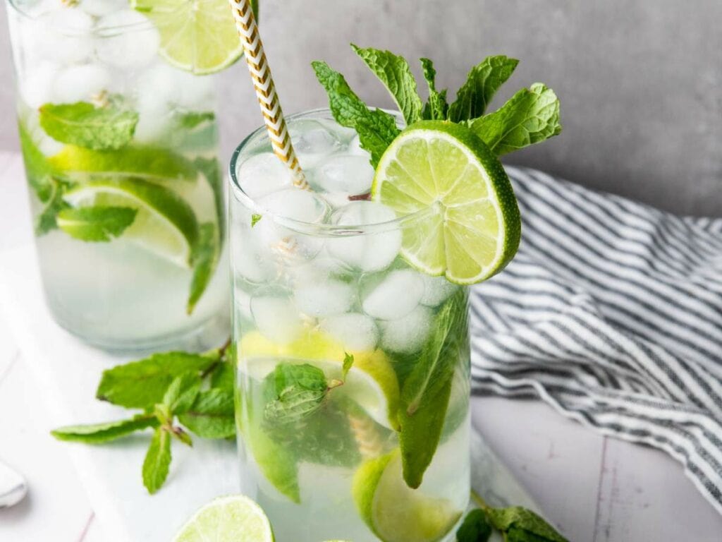A glass of mojito with ice, lime slices, fresh mint, and a striped straw sits on a white surface next to a striped cloth.