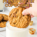 A hand dips a peanut butter cup cookie into a glass of milk, with more cookies and mini peanut butter cups in the background.