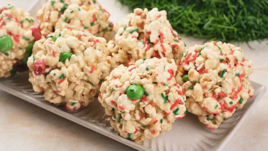 Christmas popcorn balls with colorful red, green, and white sprinkles and candy pieces are arranged on a rectangular white plate.