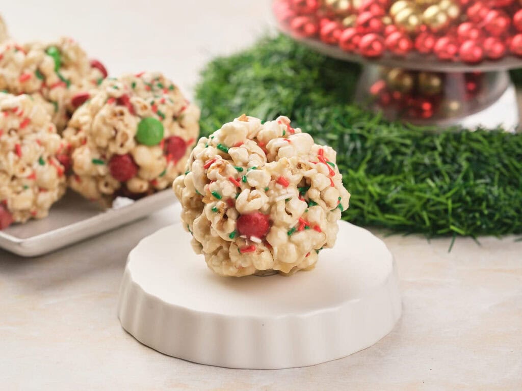 A popcorn ball with red and green candies and sprinkles sits on a white stand, with more popcorn balls and holiday decorations in the background.