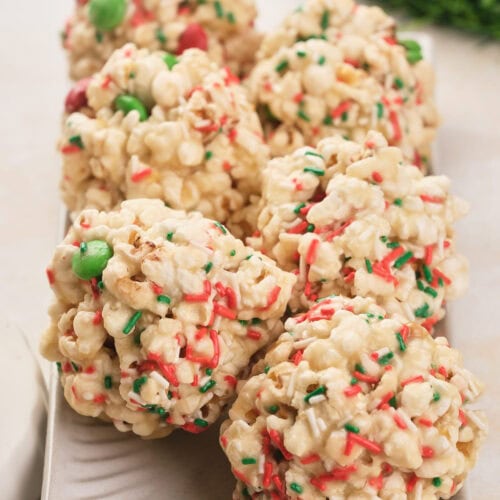 Six popcorn balls decorated with red and green sprinkles and candies are arranged on a rectangular white plate.