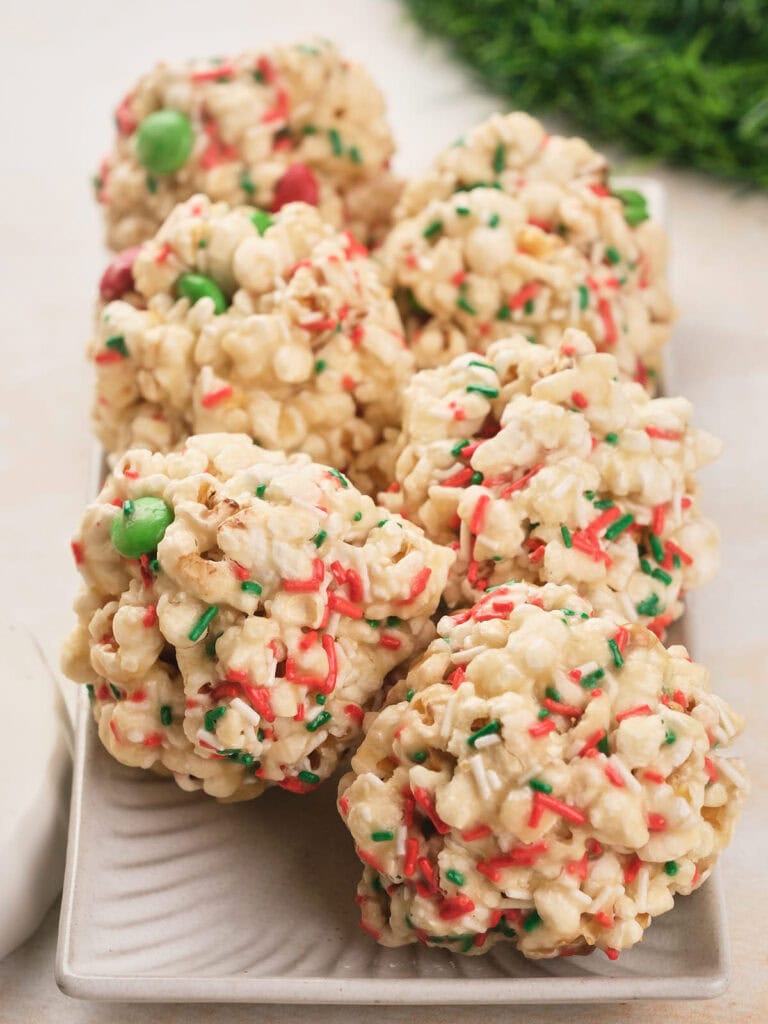 Six popcorn balls decorated with red and green sprinkles and candies are arranged on a rectangular white plate.