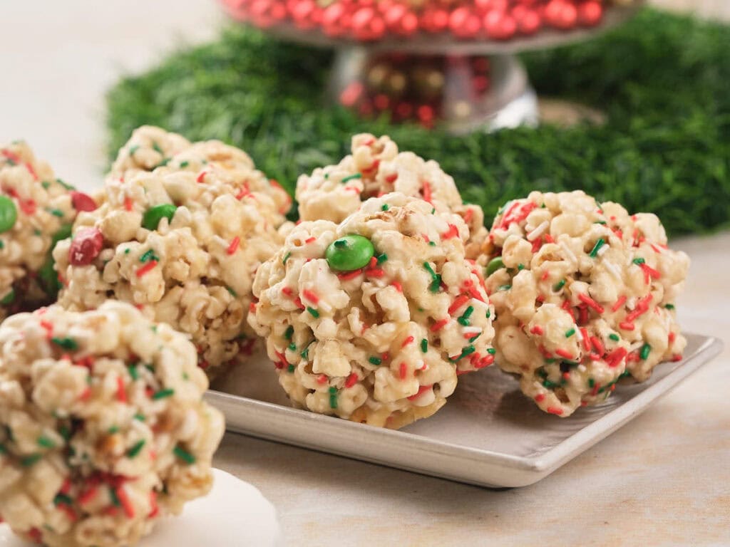 Popcorn balls with red and green sprinkles and candies are arranged on a rectangular plate with a festive background.