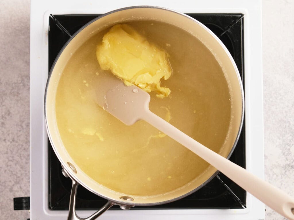 A saucepan filled with clear liquid and a large lump of yellow butter being stirred with a spatula on a stovetop.