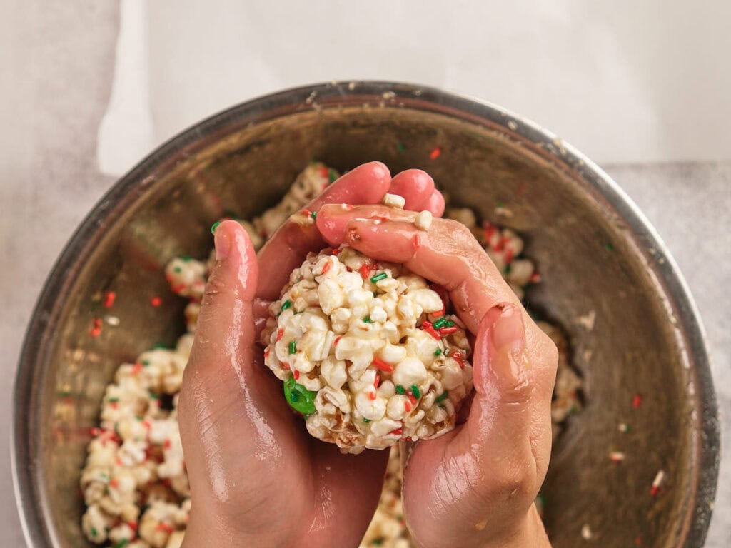 Hands shaping a ball of popcorn mixed with colorful candy sprinkles over a metal bowl filled with the popcorn mixture.