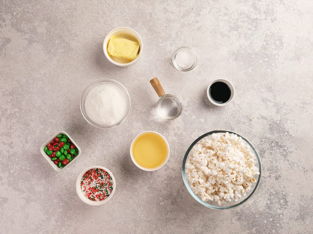 A flat lay of popcorn, butter, sugar, condensed milk, vanilla, water, sprinkles, and red and green candies in bowls on a light surface.