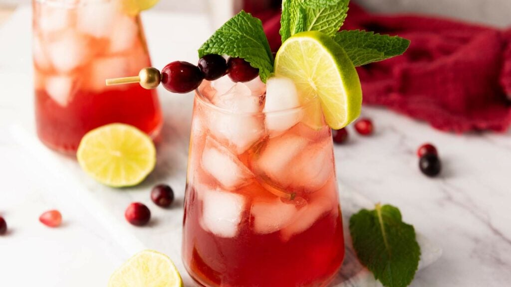 A glass of iced cranberry mocktail garnished with a lime slice, cranberries on a skewer, and fresh mint, sits on a marble surface.