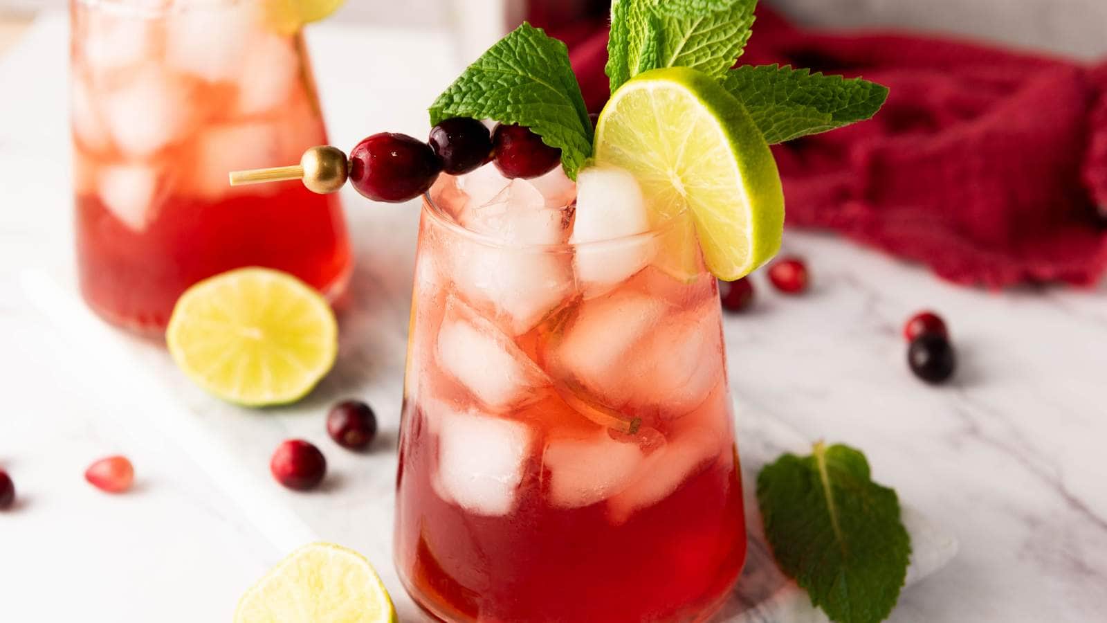 A glass of iced cranberry mocktail garnished with a lime slice, cranberries on a skewer, and fresh mint, sits on a marble surface.