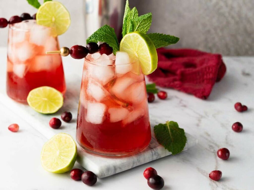 Two cranberry mocktails garnished with lime slices, mint sprigs, and skewered cranberries sit on a marble surface.