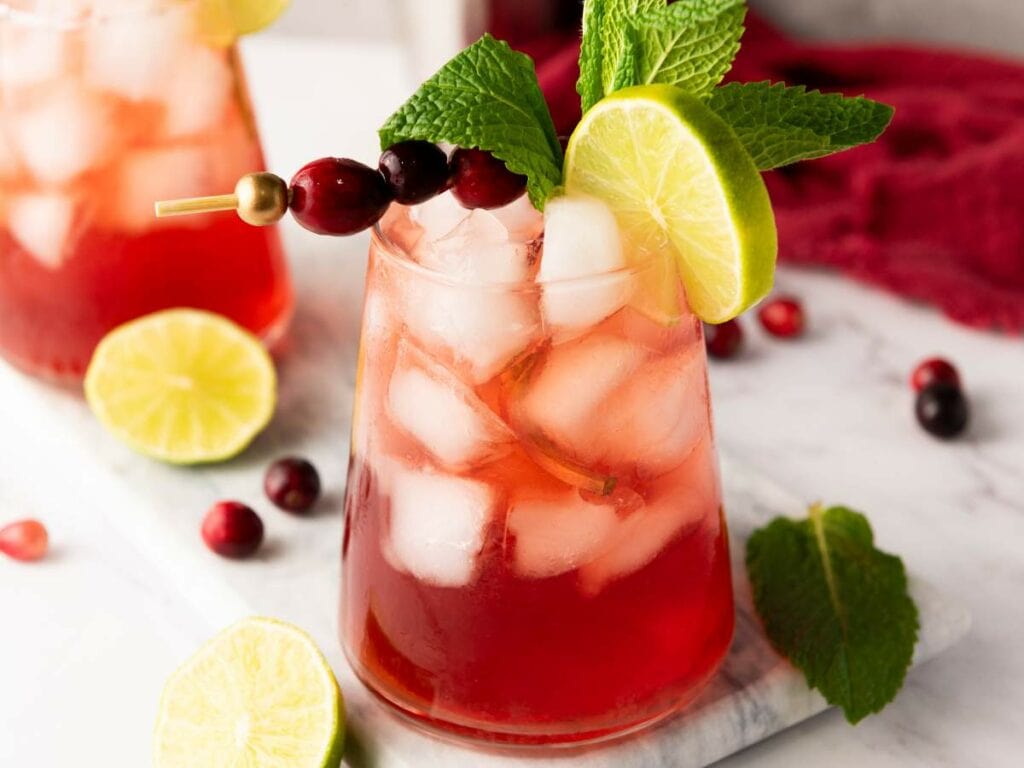 A glass of cranberry mocktail garnished with a lime slice, fresh mint, and cranberries on a skewer, with ice cubes and lime wedges nearby.