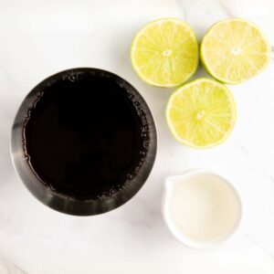 A metal cup filled with a dark liquid, three halves of lime, and a small pitcher of white liquid on a white surface.