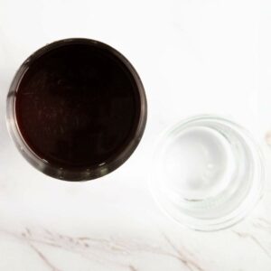 A top-down view of a glass filled with a dark liquid next to an empty clear glass, both on a white surface.