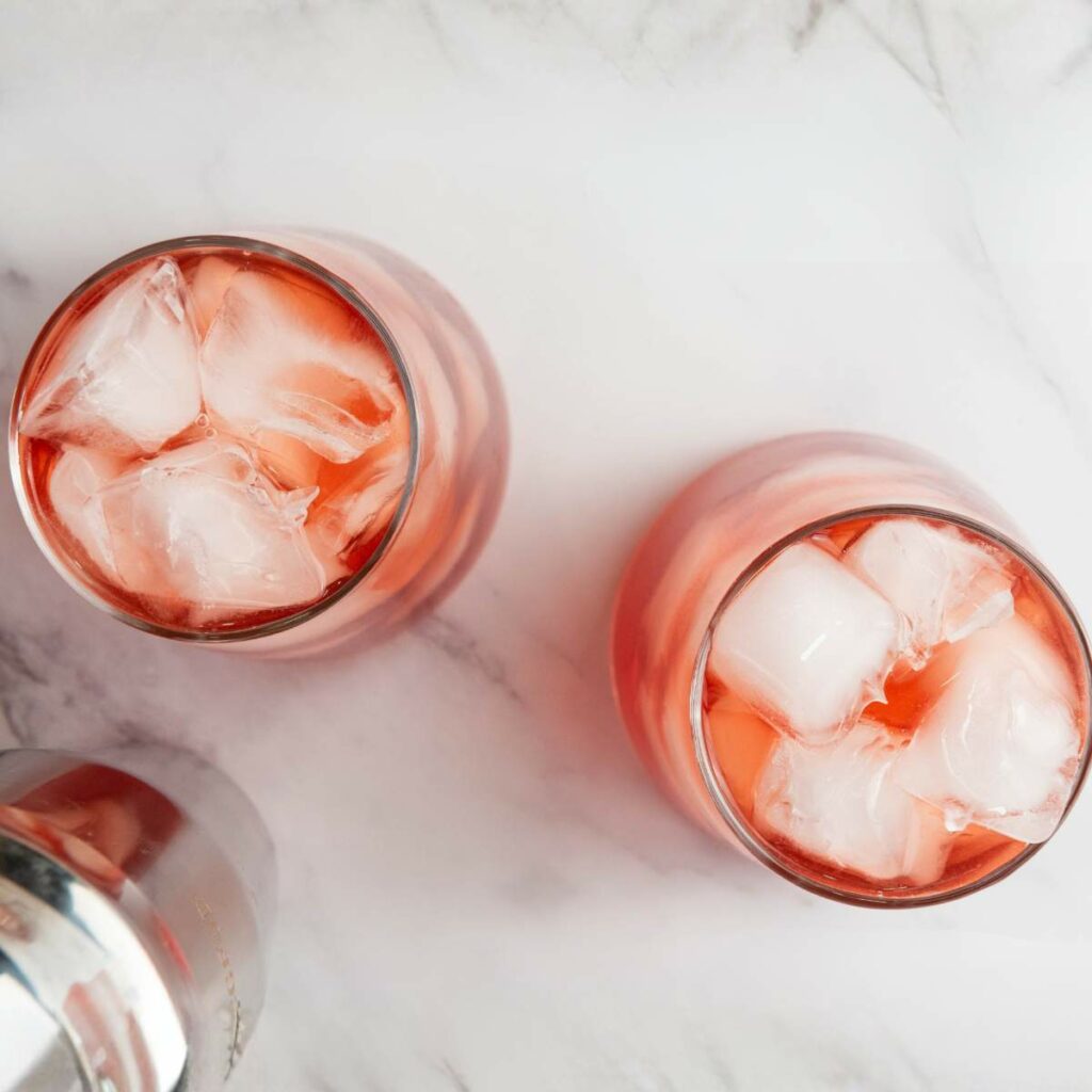 Two glasses filled with a pink iced beverage sit on a white marble surface, with a metal cocktail shaker partially visible in the corner.