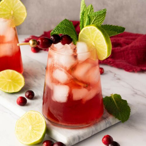 A glass of red iced cranberry mocktail garnished with a lime slice, mint leaves, and cranberries on a marble surface.