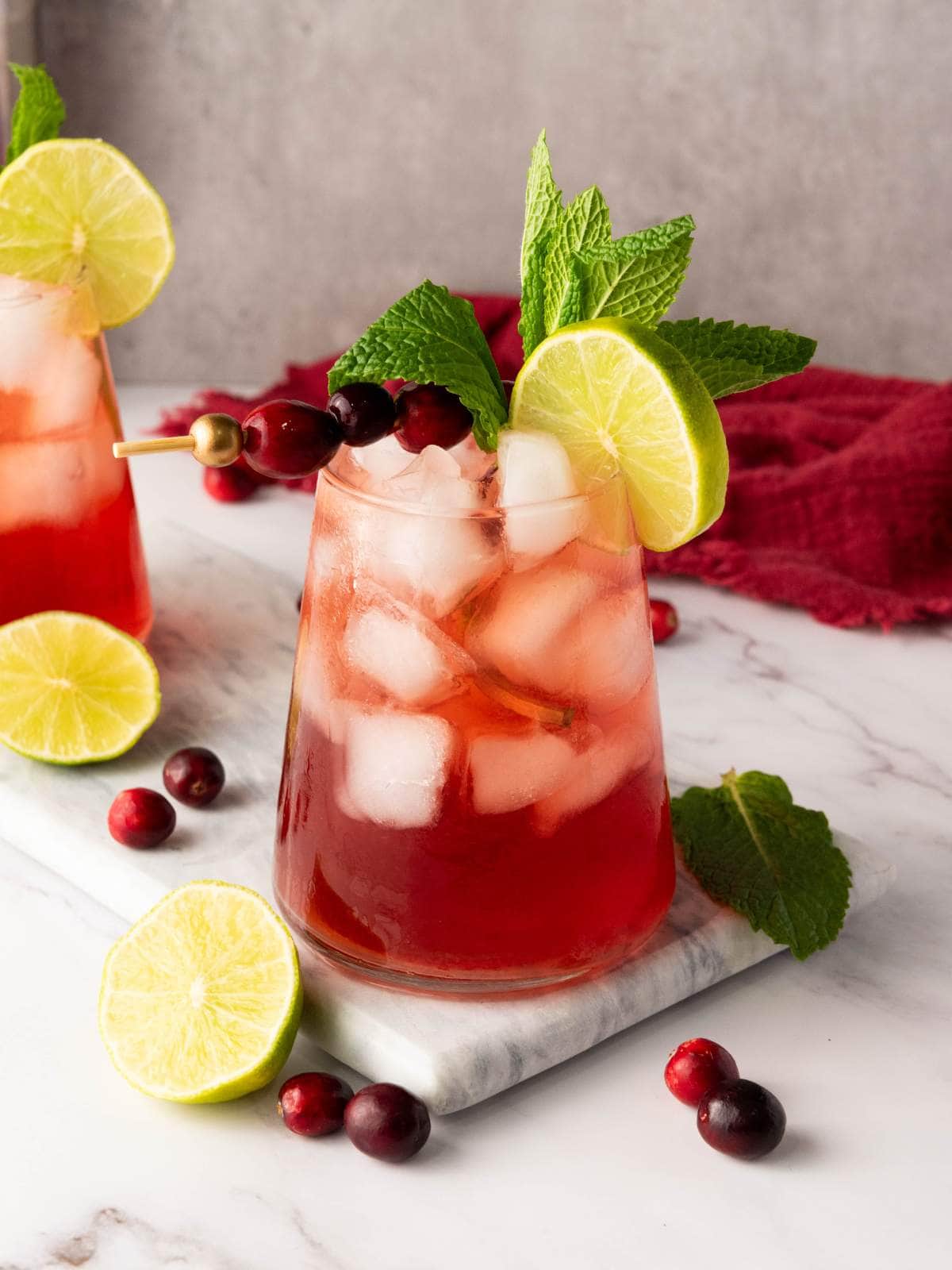 A glass of red iced cranberry mocktail garnished with a lime slice, mint leaves, and cranberries on a marble surface.