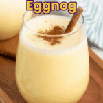 A glass of eggnog topped with ground cinnamon and a cinnamon stick, placed on a wooden tray next to another glass.