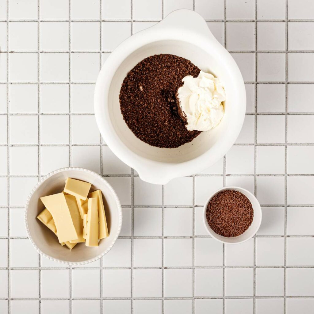 Three bowls on a tiled surface: one with chocolate crumbs and cream cheese, one with white chocolate pieces, and one with chocolate sprinkles.