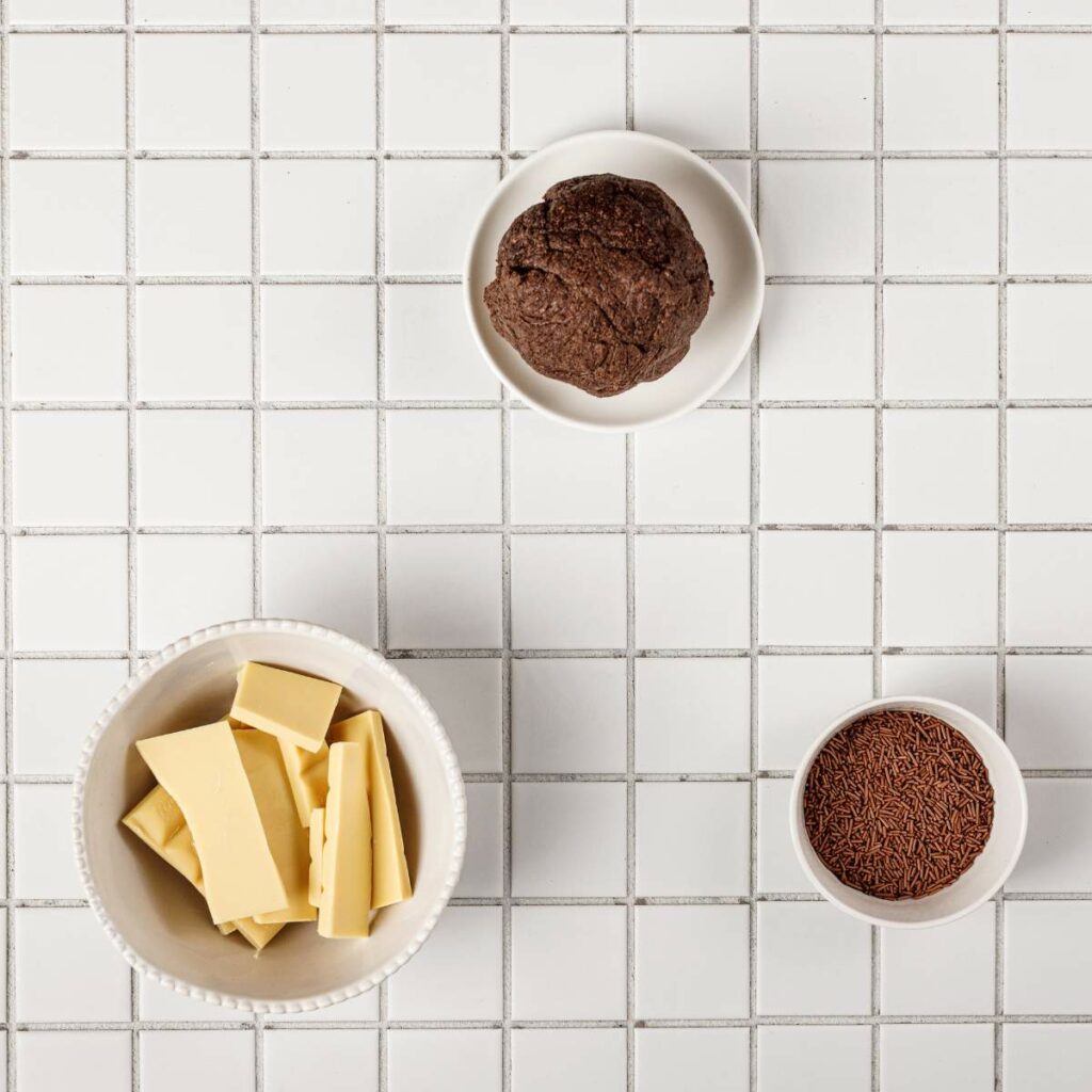 Three bowls on a white tiled surface: one with chocolate dough, one with chocolate sprinkles, and one with white chocolate pieces.