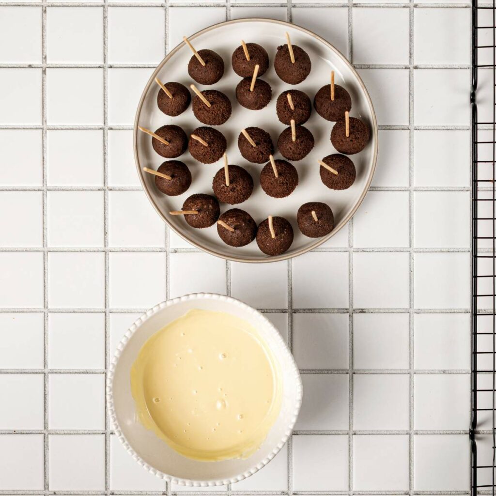 A plate of round chocolate cake balls with toothpicks next to a bowl of white melted coating on a white tiled surface.