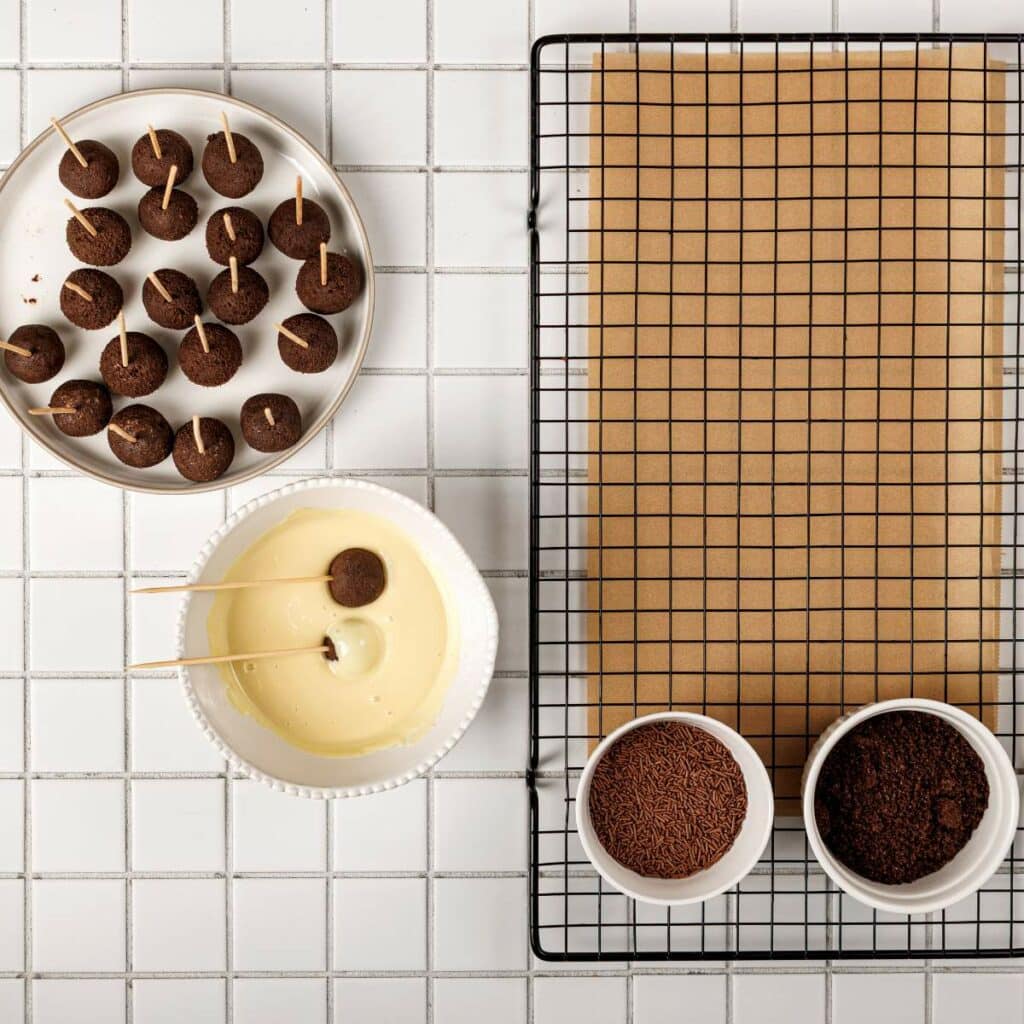 Overhead view of cake pops on sticks, a bowl of white coating, a cooling rack with parchment paper.