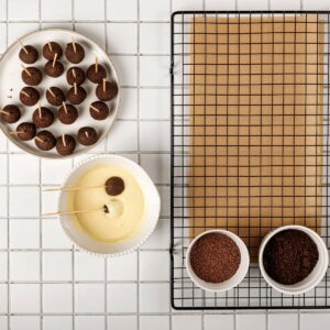 Overhead view of cake pops on sticks, a bowl of white coating, a cooling rack with parchment paper.