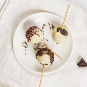 Three cake pops coated in white chocolate with chocolate sprinkles, served on a white plate with a beige napkin underneath.