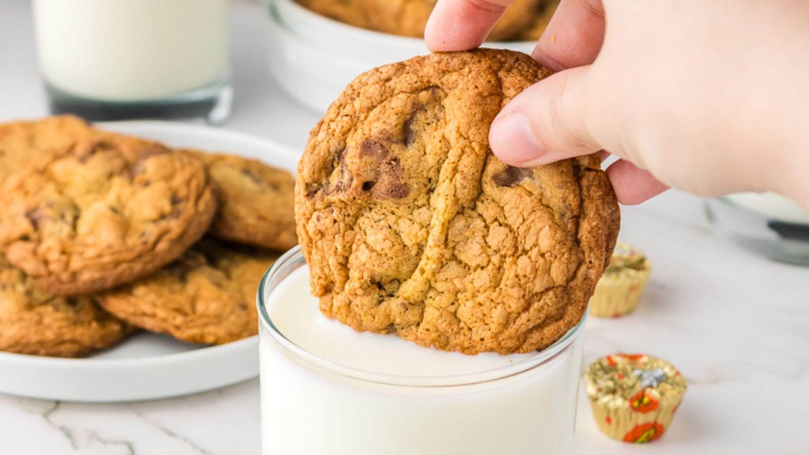 A hand dips a peanut butter cup cookie into a glass of milk, with more cookies and milk in the background on a white surface.
