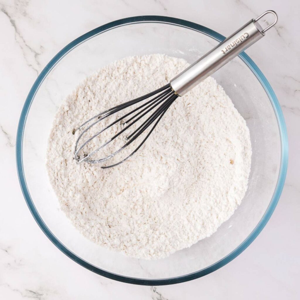 A glass bowl filled with flour mixture and a metal whisk resting on top, placed on a white marble surface.