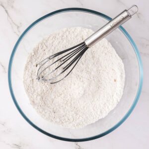 A glass bowl filled with flour mixture and a metal whisk resting on top, placed on a white marble surface.