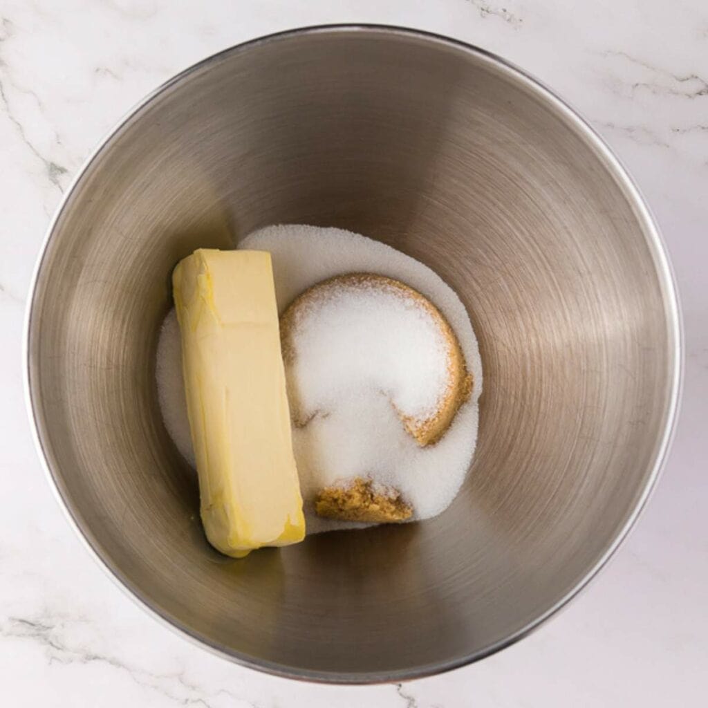 A stick of butter, white sugar, and brown sugar in a large stainless steel mixing bowl on a marble countertop.