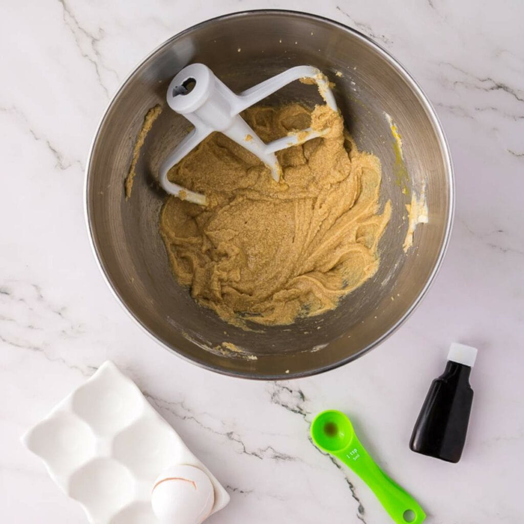 A mixing bowl with creamed butter and sugar, a paddle attachment, a cracked egg in a ceramic tray, and a green measuring spoon.